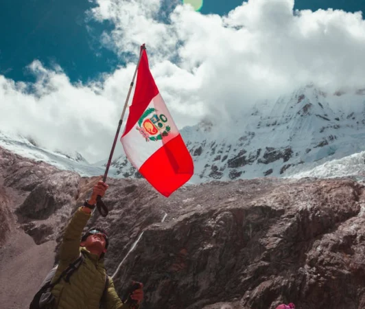 Hiker proudly waves the Peruvian flag against a stunning mountainous background with snow-capped peaks.